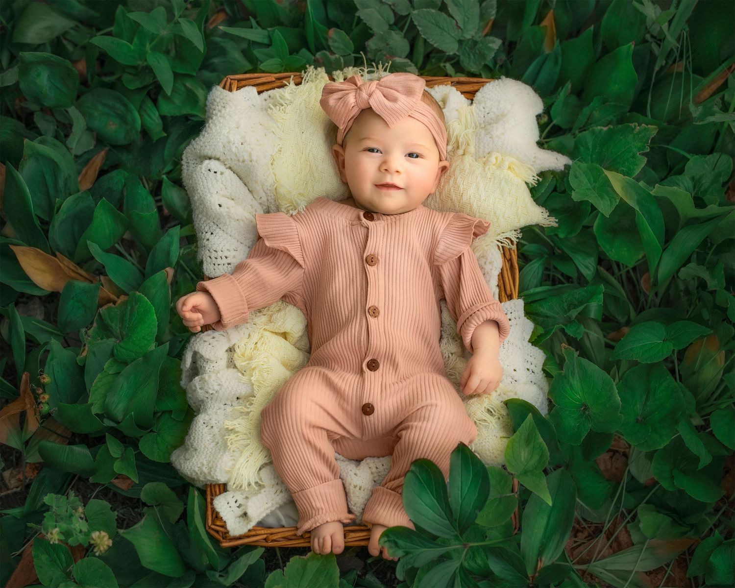 Baby in pink knit romper lying on white textured basket of vintage blankets surrounded by greenery in a garden.