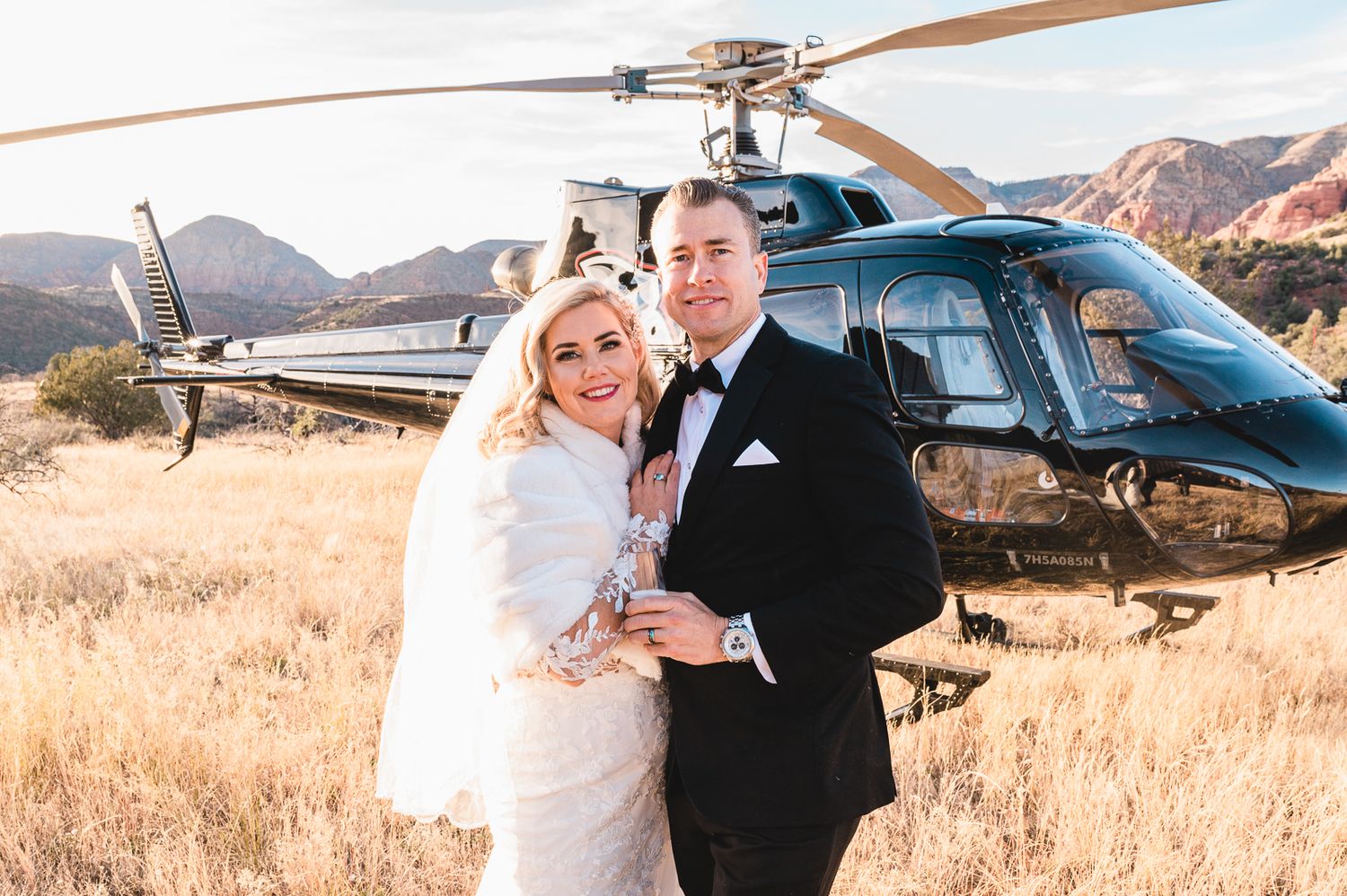 Wedding couple poses in front of helicopter in desert mountain landscape.