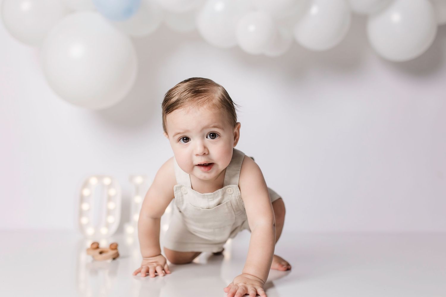 Baby in white outfit crawls on floor under white balloon arch with decorative lights.