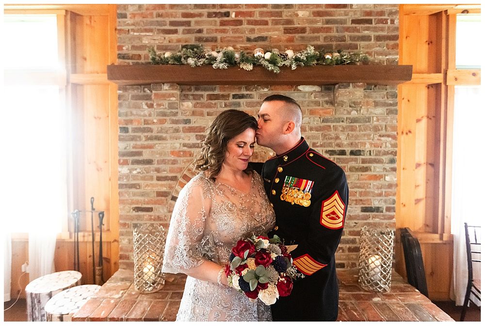 Marine groom kisses bride on cheek as she holds red and white bouquet in front of brick fireplace.