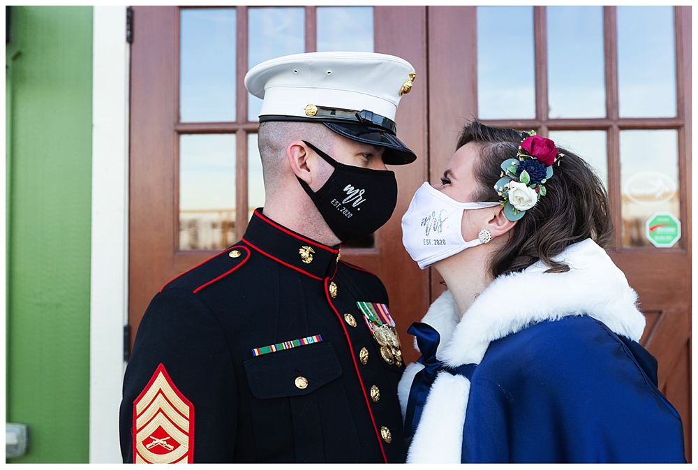 Marine in dress uniform and bride in blue cape share a masked kiss outdoors during COVID wedding.