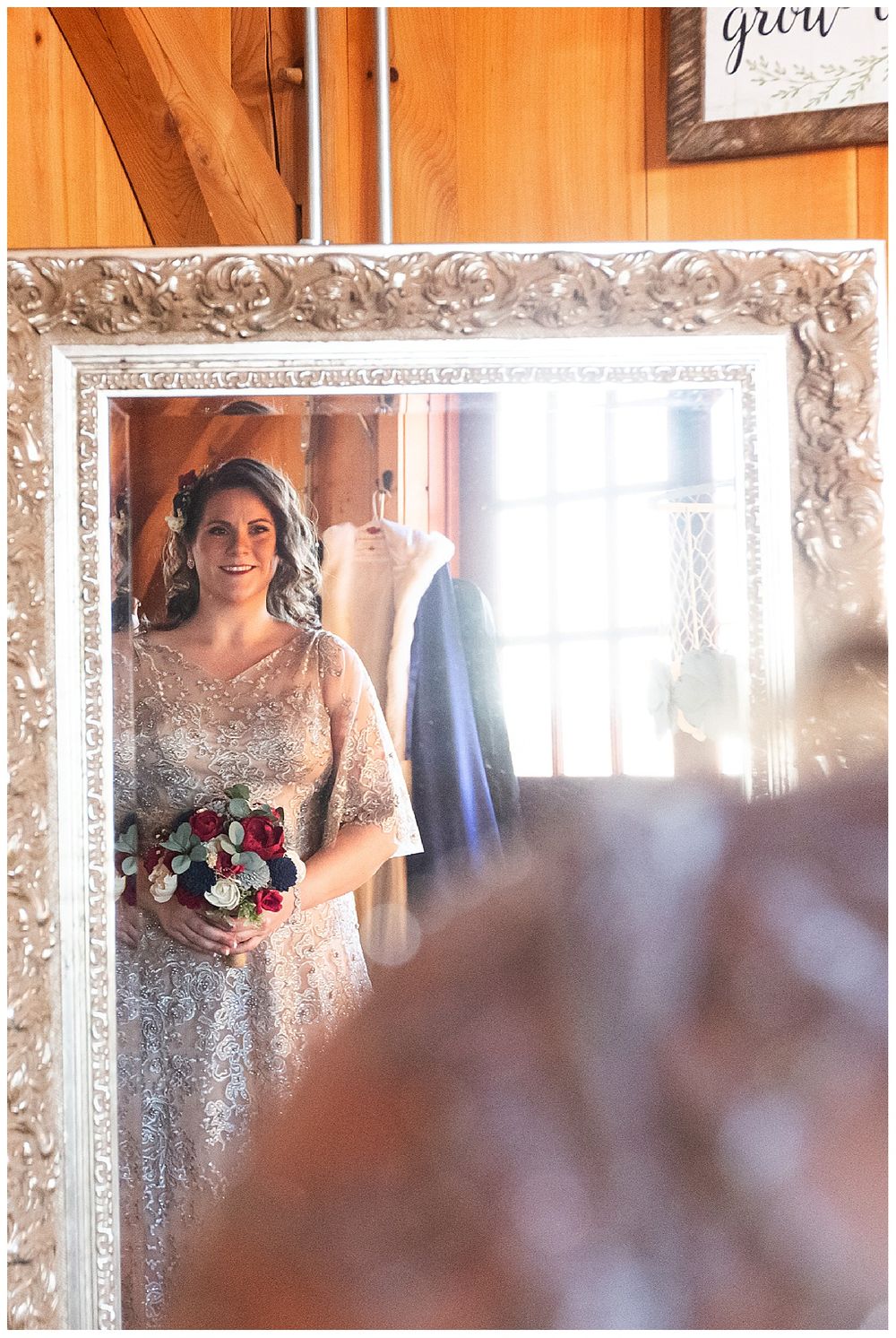 Smiling bride holding red and white bouquet reflected in ornate silver mirror in rustic bridal suite.