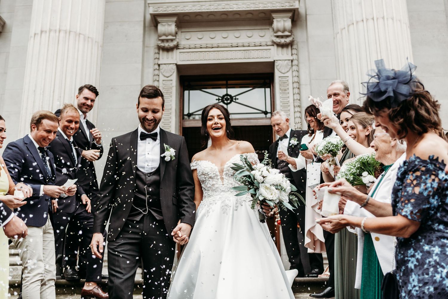 Wedding party celebrates with confetti as newlyweds exit historic building with columns.