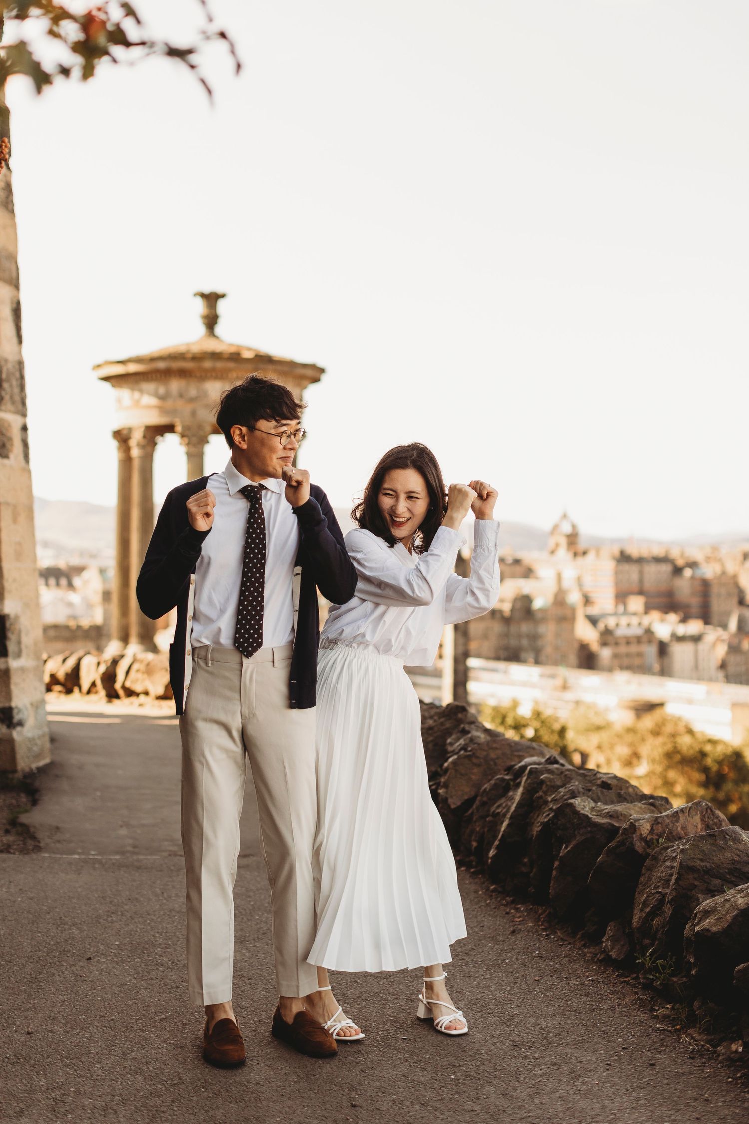 A couple in elegant white and tan outfits poses playfully at a scenic viewpoint.