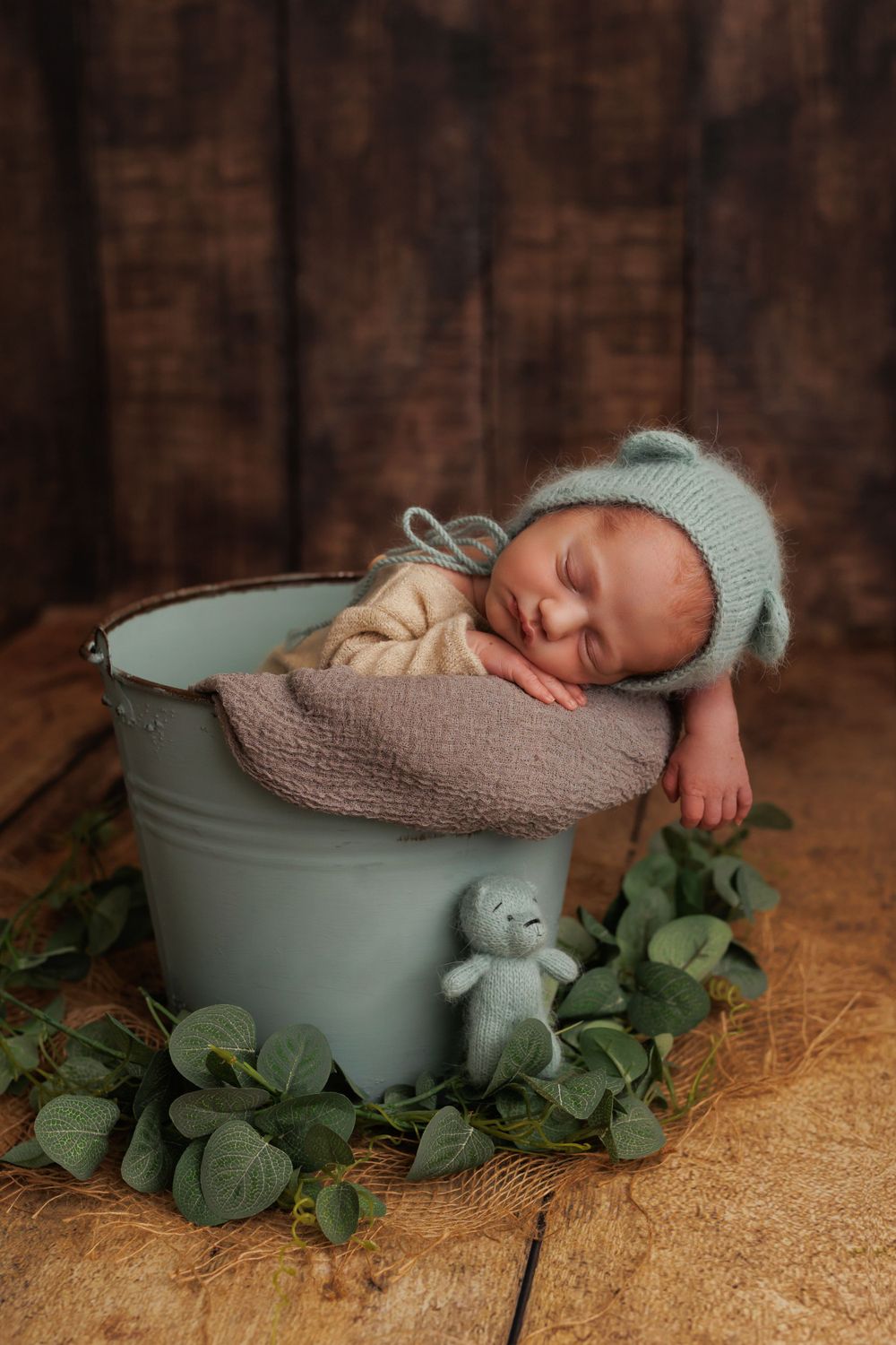 Sleeping newborn in mint green hat nestled in bucket with eucalyptus leaves and teddy bear.