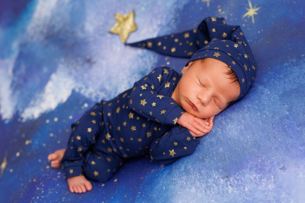 Sleeping newborn in starry blue outfit against dreamy night sky backdrop.
