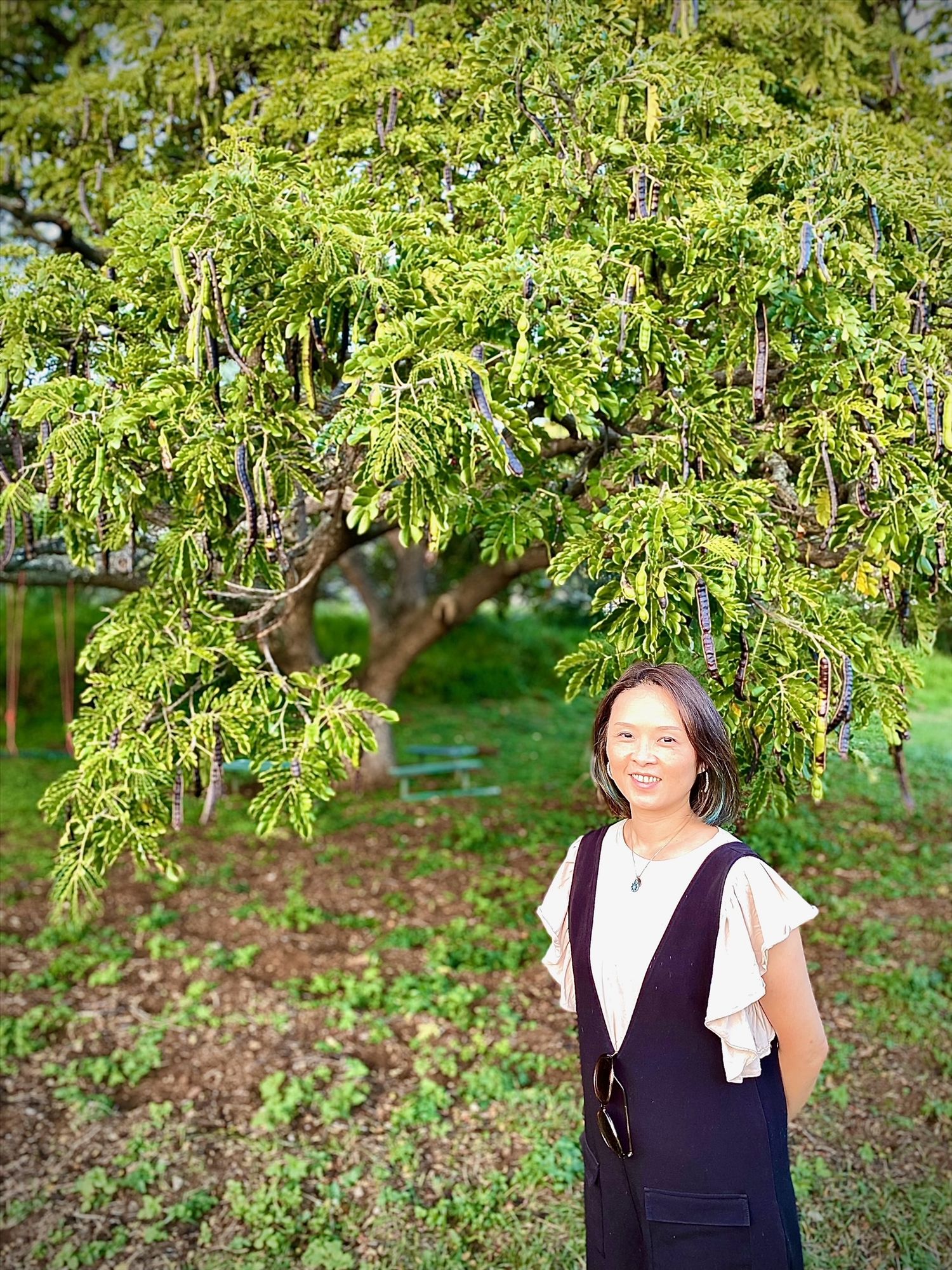 Smiling woman in black overalls stands beneath a large lush green tree in a tropical park setting.
ウェディングプランナー