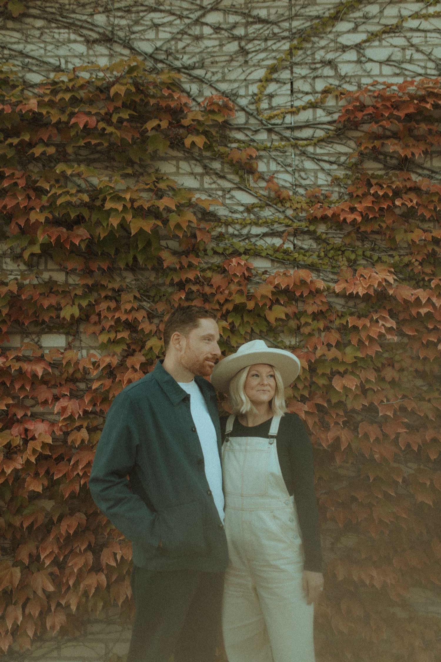 A couple poses against a wall covered in colorful autumn ivy wearing casual fall attire.
