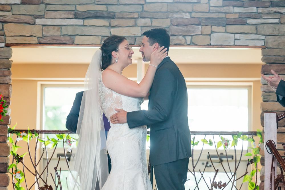 A romantic moment between newlyweds sharing their first kiss in front of a stone wall and window backdrop.