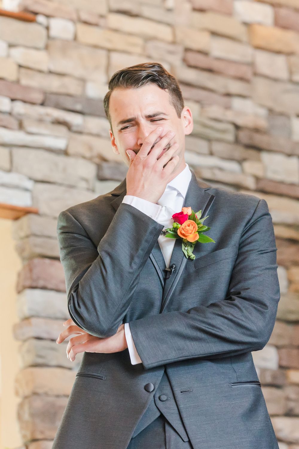 A groom in a gray suit with an orange boutonniere shows an emotional expression against a stone wall.