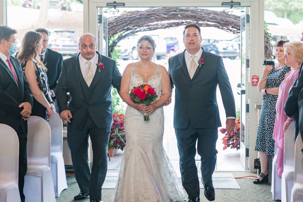 Wedding party walking down venue aisle with bride in mermaid gown holding red bouquet.