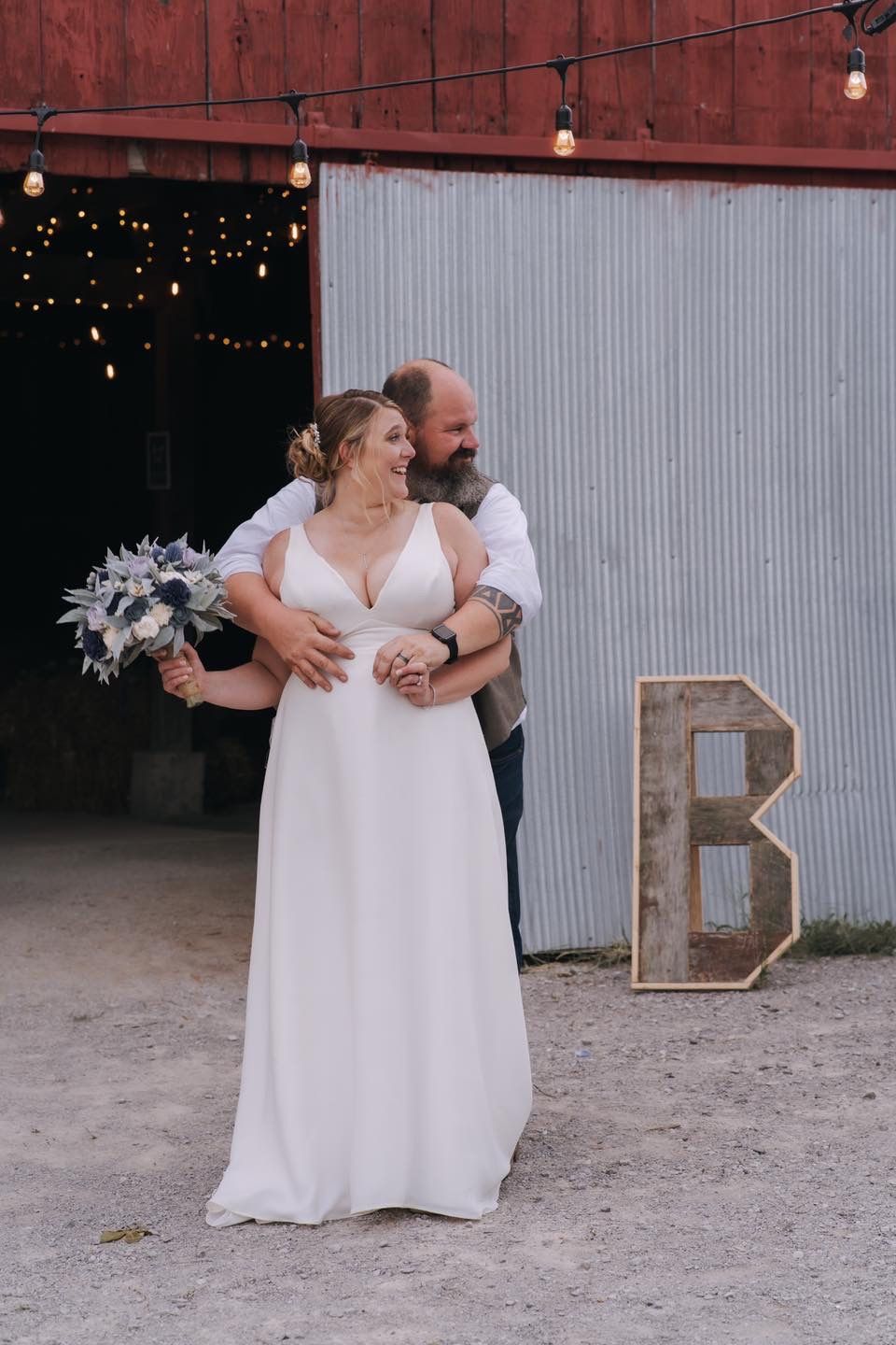 A rustic barn wedding scene with string lights and a couple sharing an intimate moment beside a metal letter B.