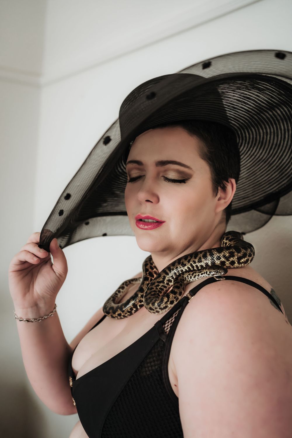 short haired woman in red lipstick and glamour makeup wearing fashion hat with snake around neck