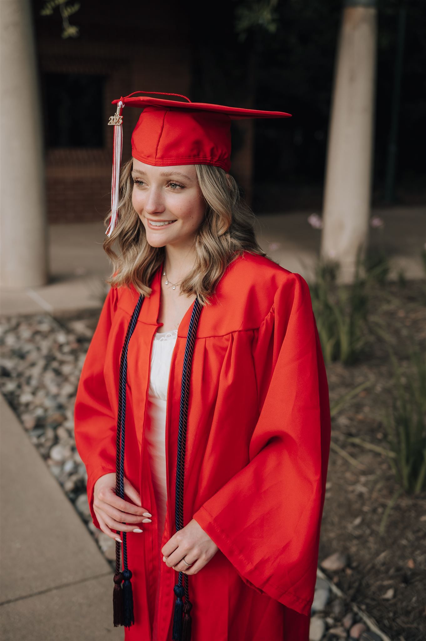 A graduate poses in traditional red cap and gown against stone columns and landscaping.