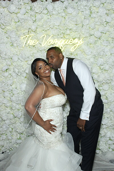 A romantic wedding photo with a white floral wall backdrop and neon sign reading 'The Varneys'.