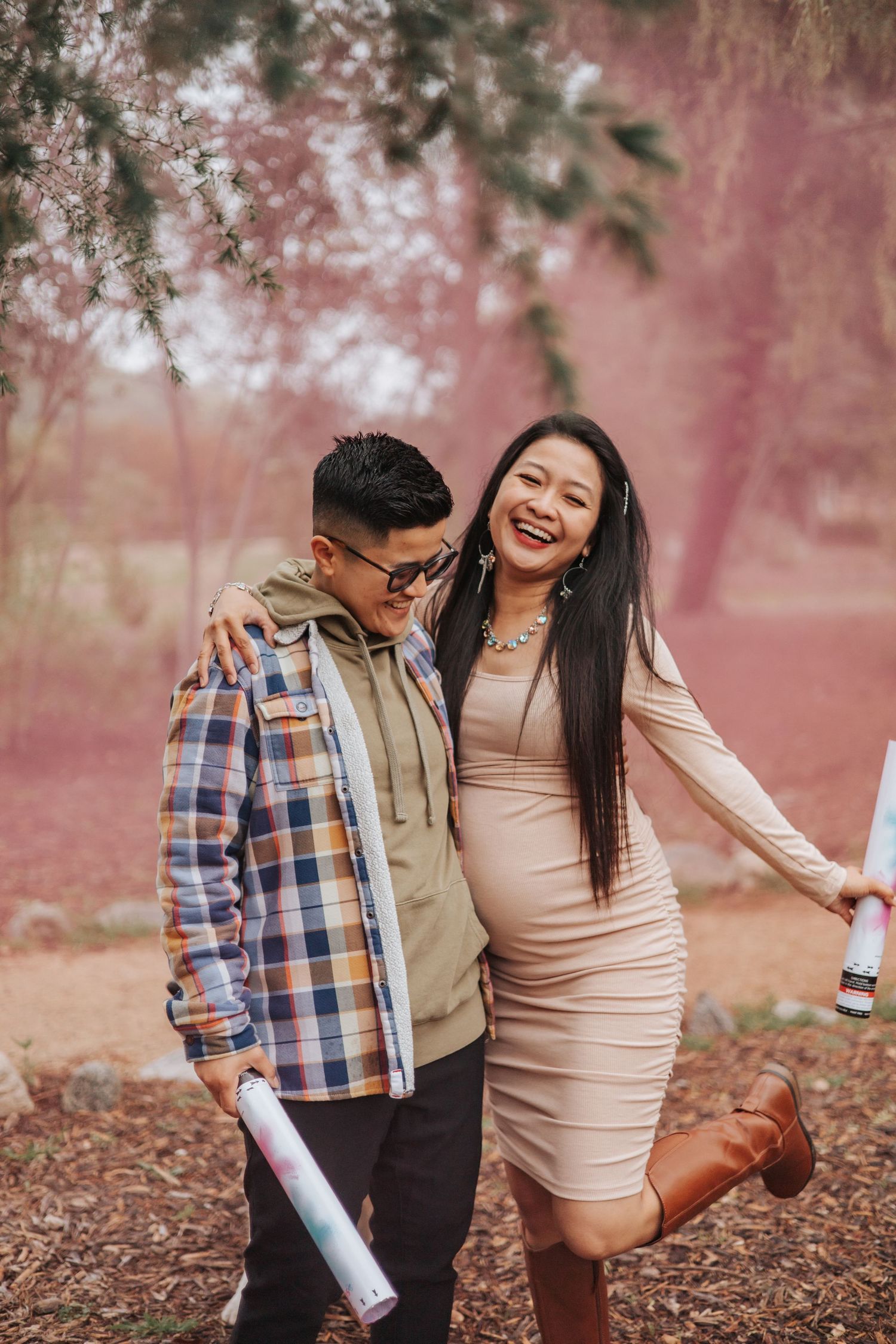 A joyful couple posing with pink smoke effects in a wooded area.