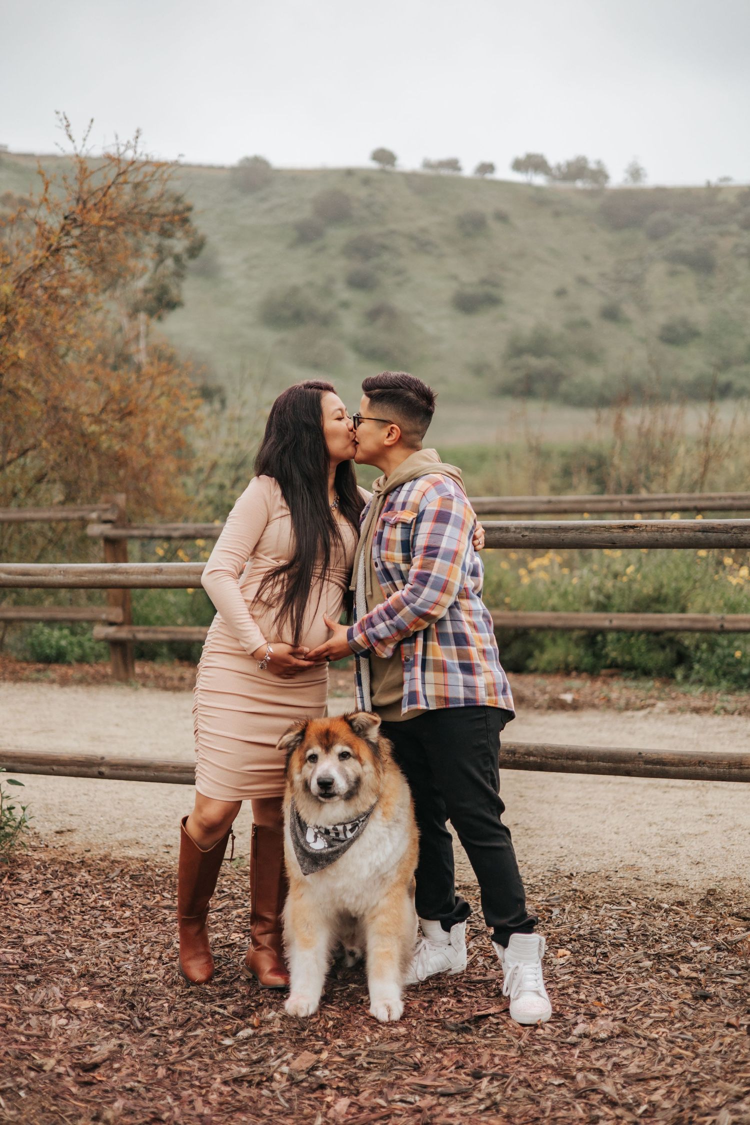 A couple shares a romantic kiss while their fluffy dog sits patiently on a dirt path with rolling hills in the background.