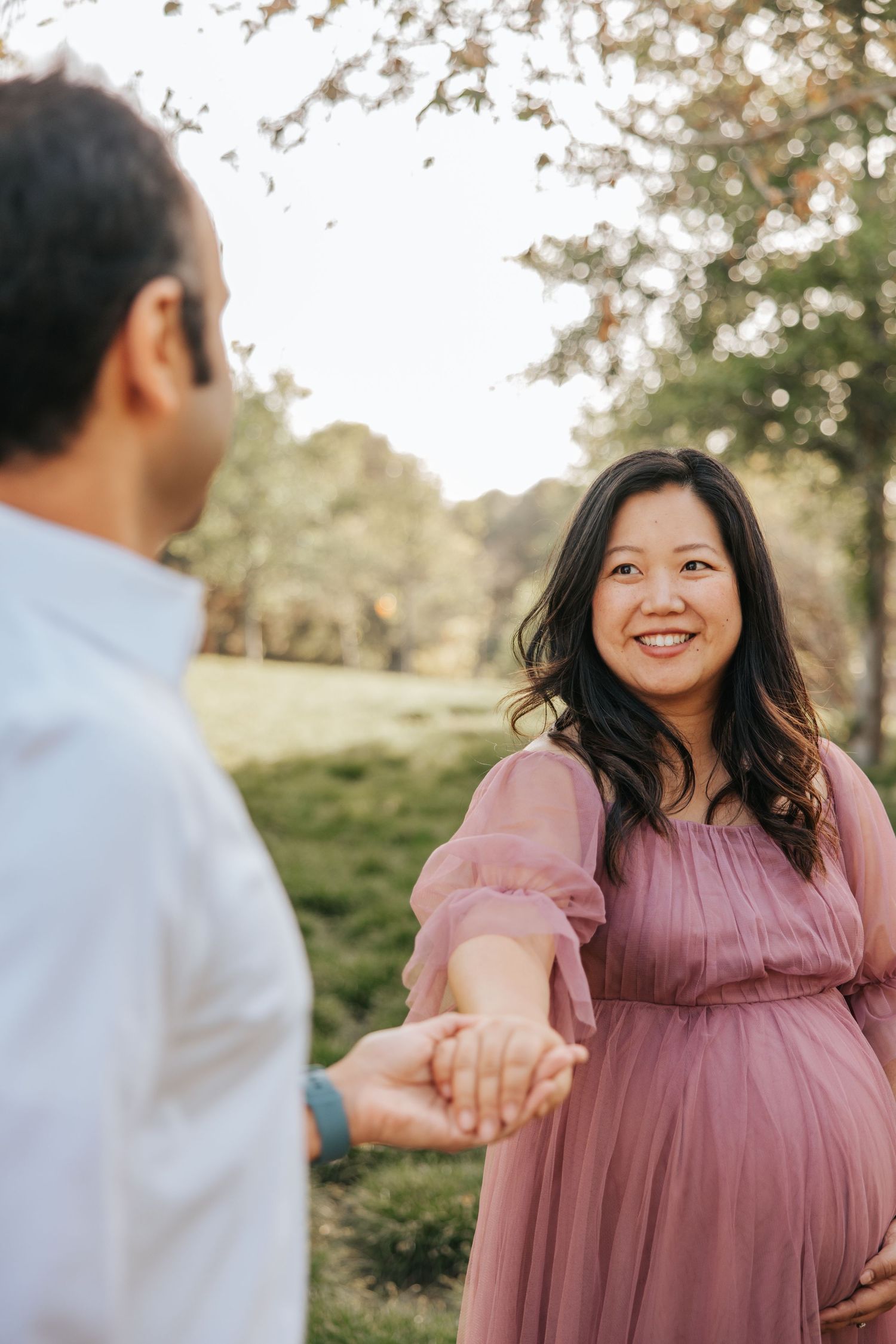 A couple holding hands outdoors as the sun sets through trees in the background.