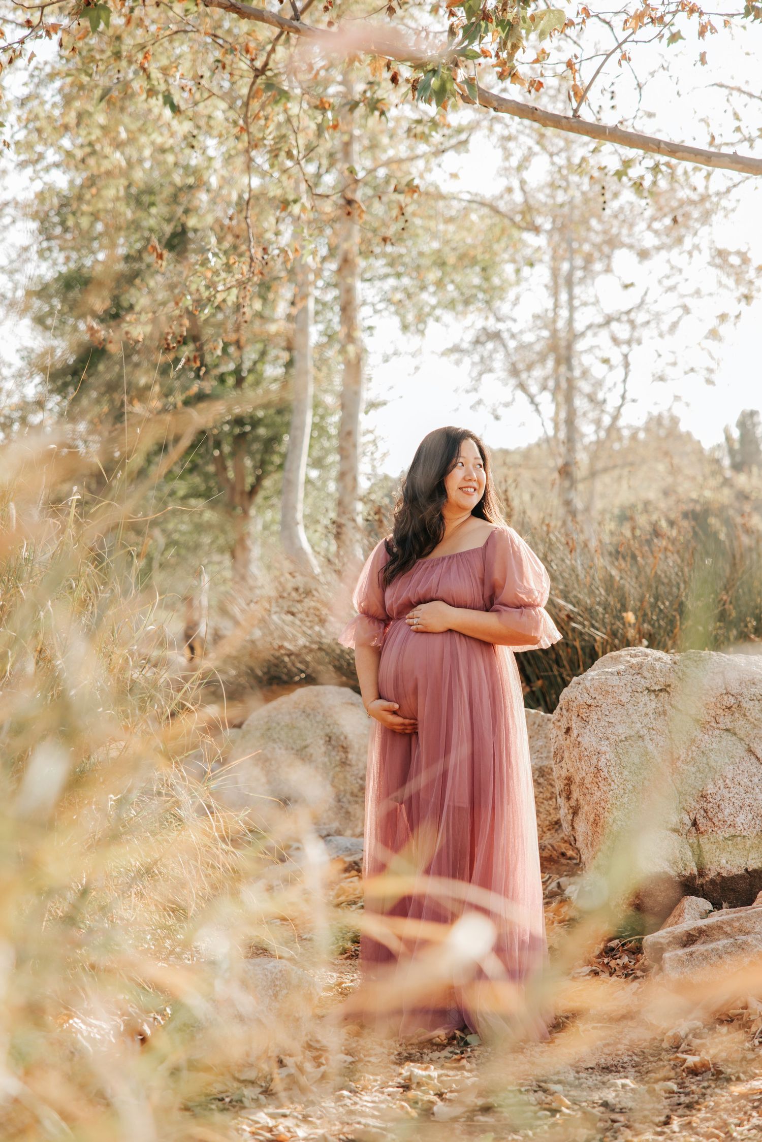 A person in a flowing pink maternity dress poses against a dreamy natural backdrop.