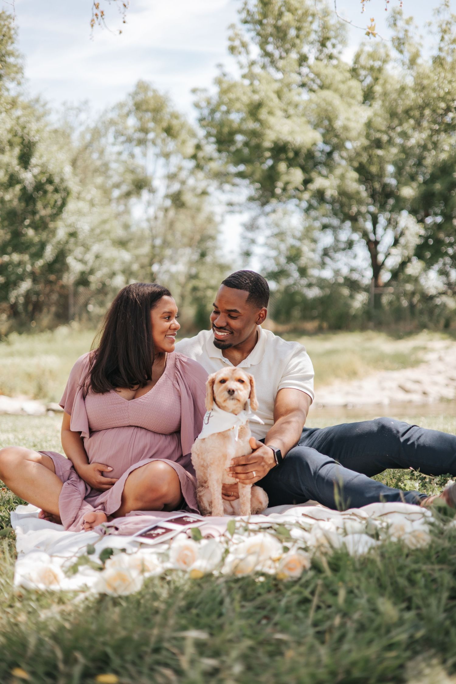 A couple relaxes on a picnic blanket with their small dog in a sunny park setting.