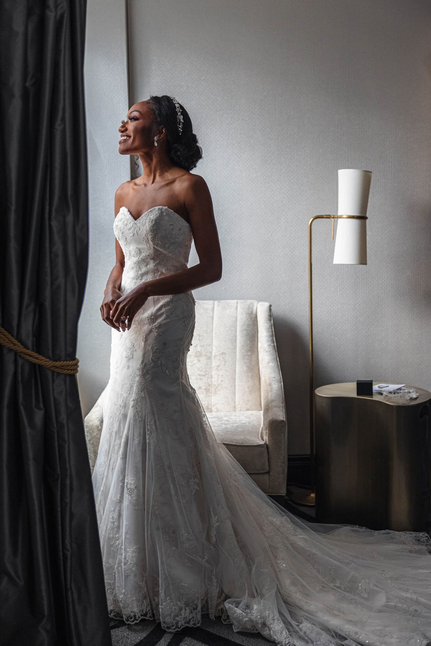 A bride in a fitted lace wedding dress stands by a modern floor lamp in an elegant room.