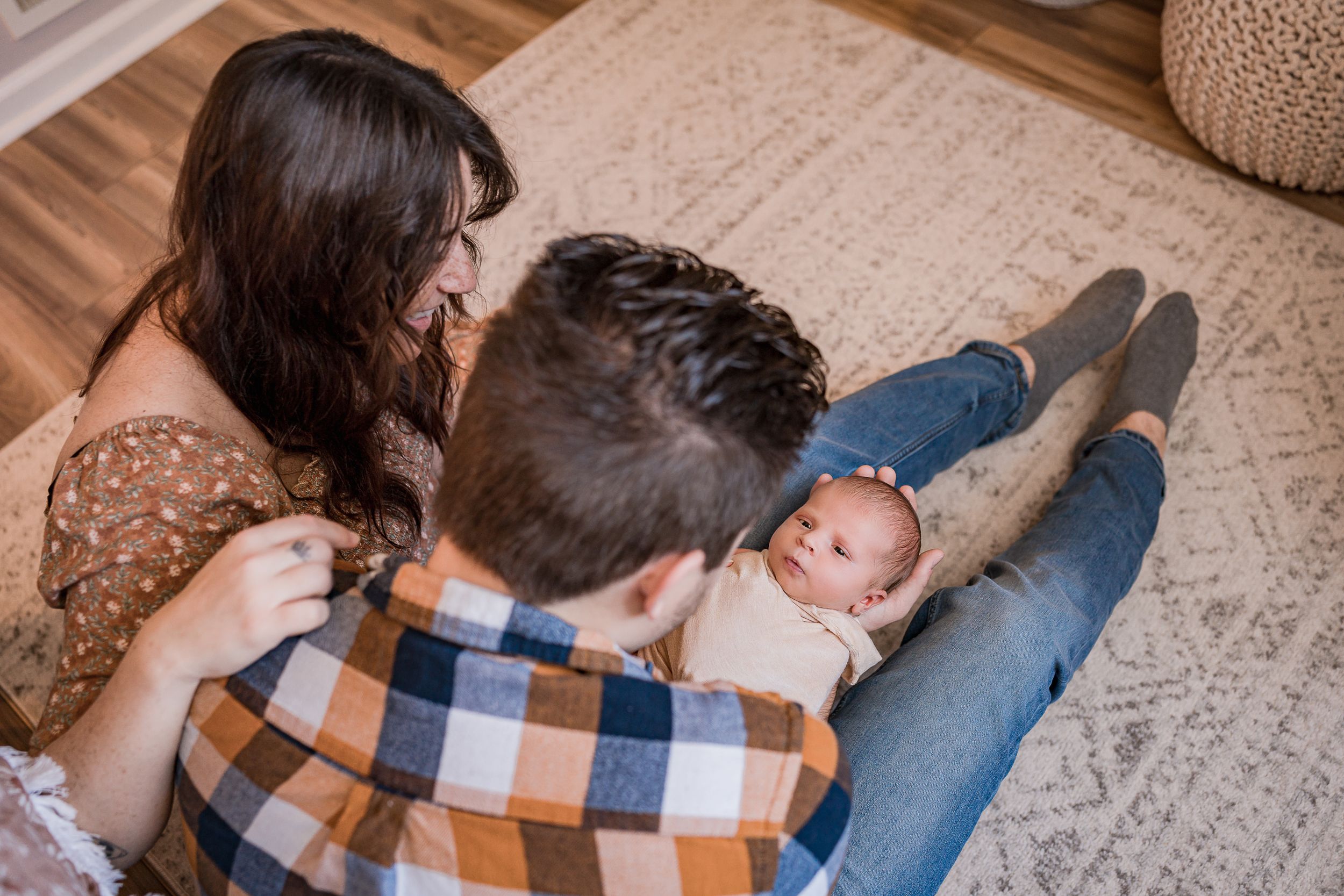 New parents snuggle with their tiny baby on a patterned carpet in a cozy living room.