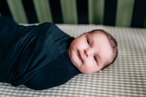 Baby in navy blue swaddle lies on striped bedding with bright eyes.