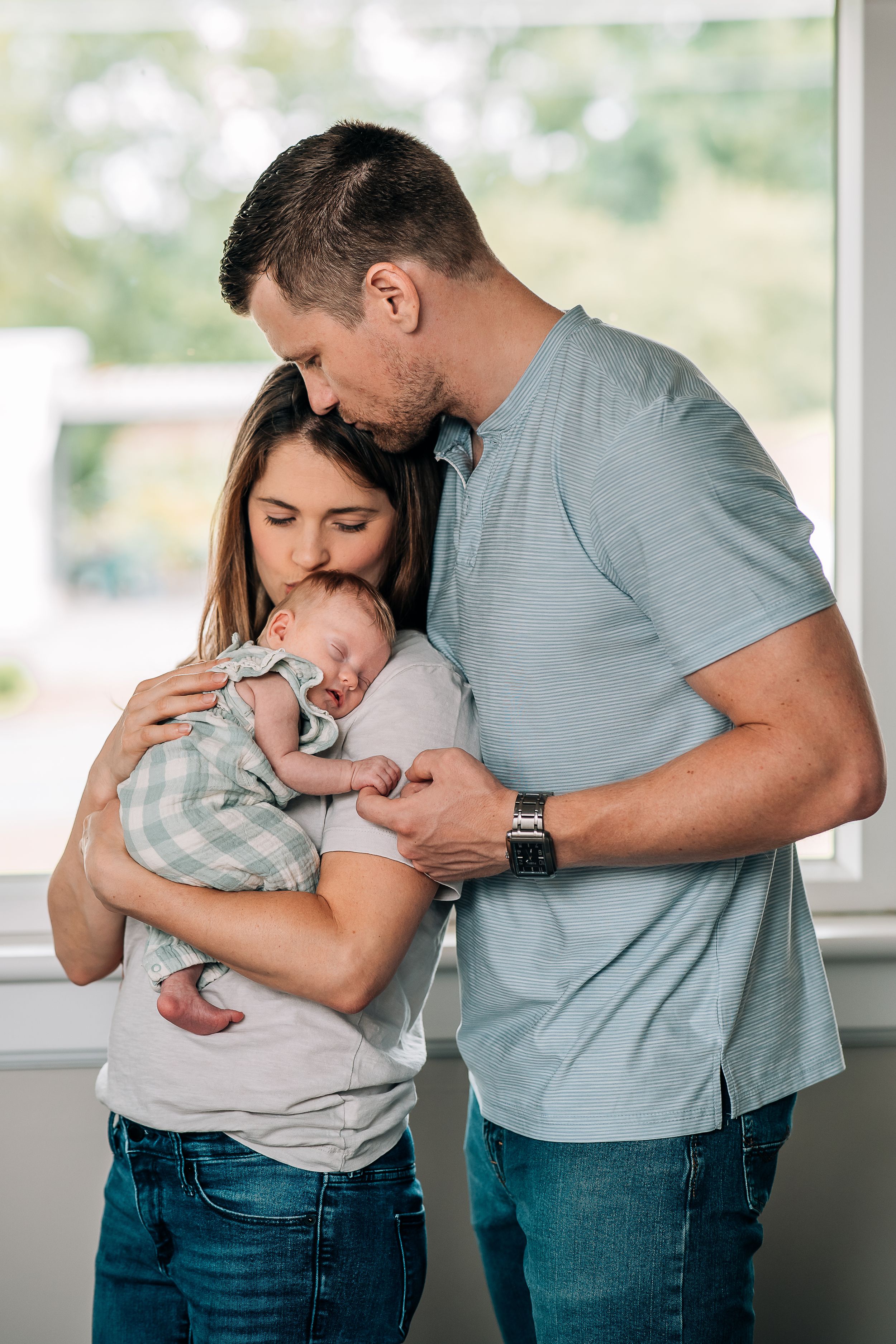 Family embraces while holding sleeping baby near bright window.