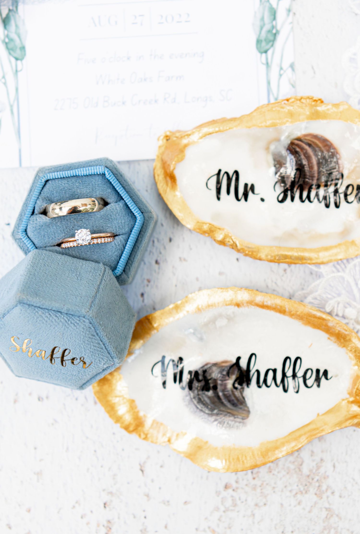 Wedding rings displayed in a blue hexagonal box next to personalized oyster shells.