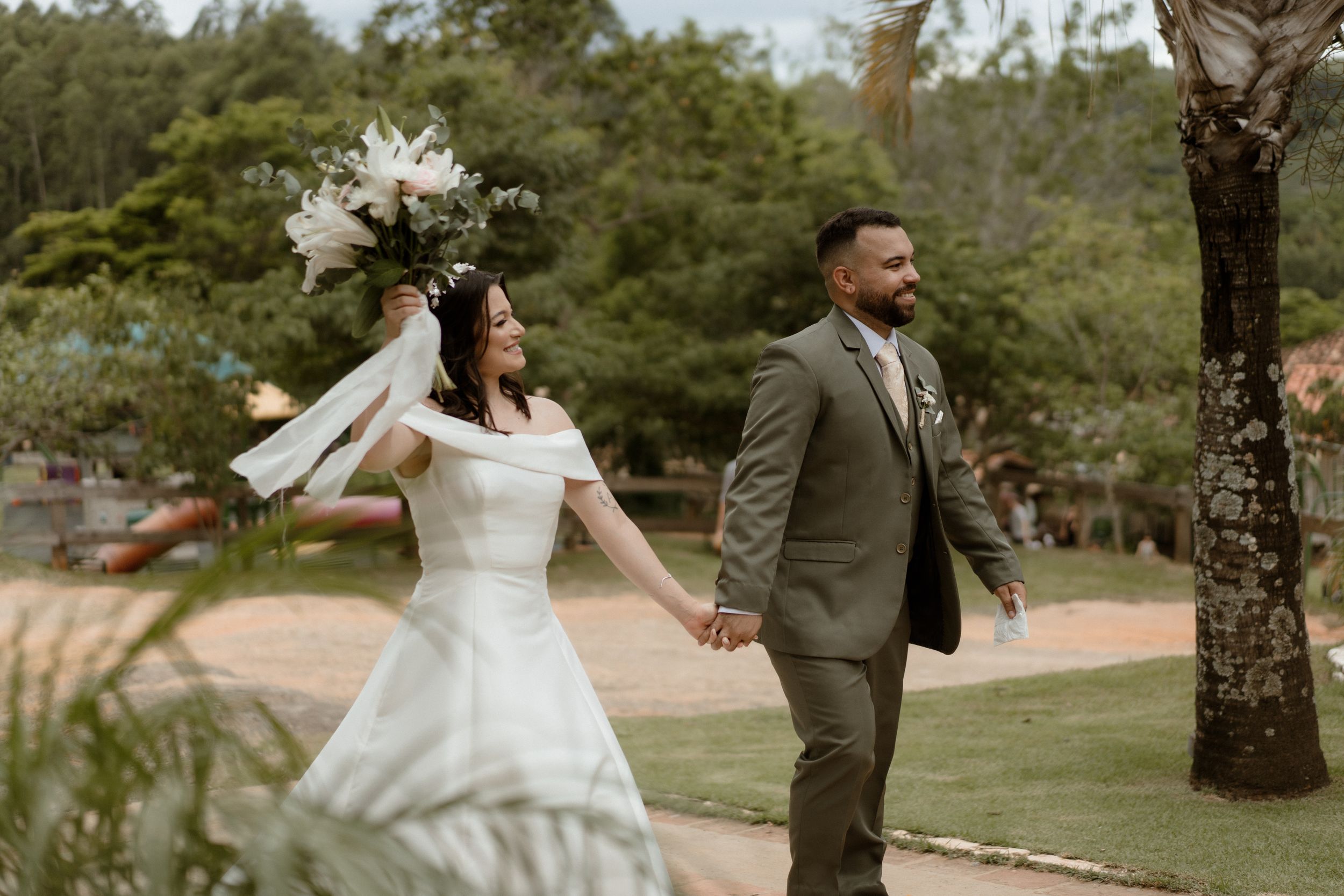 Couple walks hand in hand through garden venue with palm trees and white flowers.