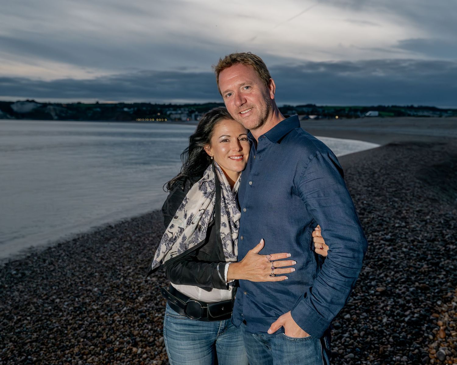 Couple poses together on pebble beach with water and cloudy sky in background.