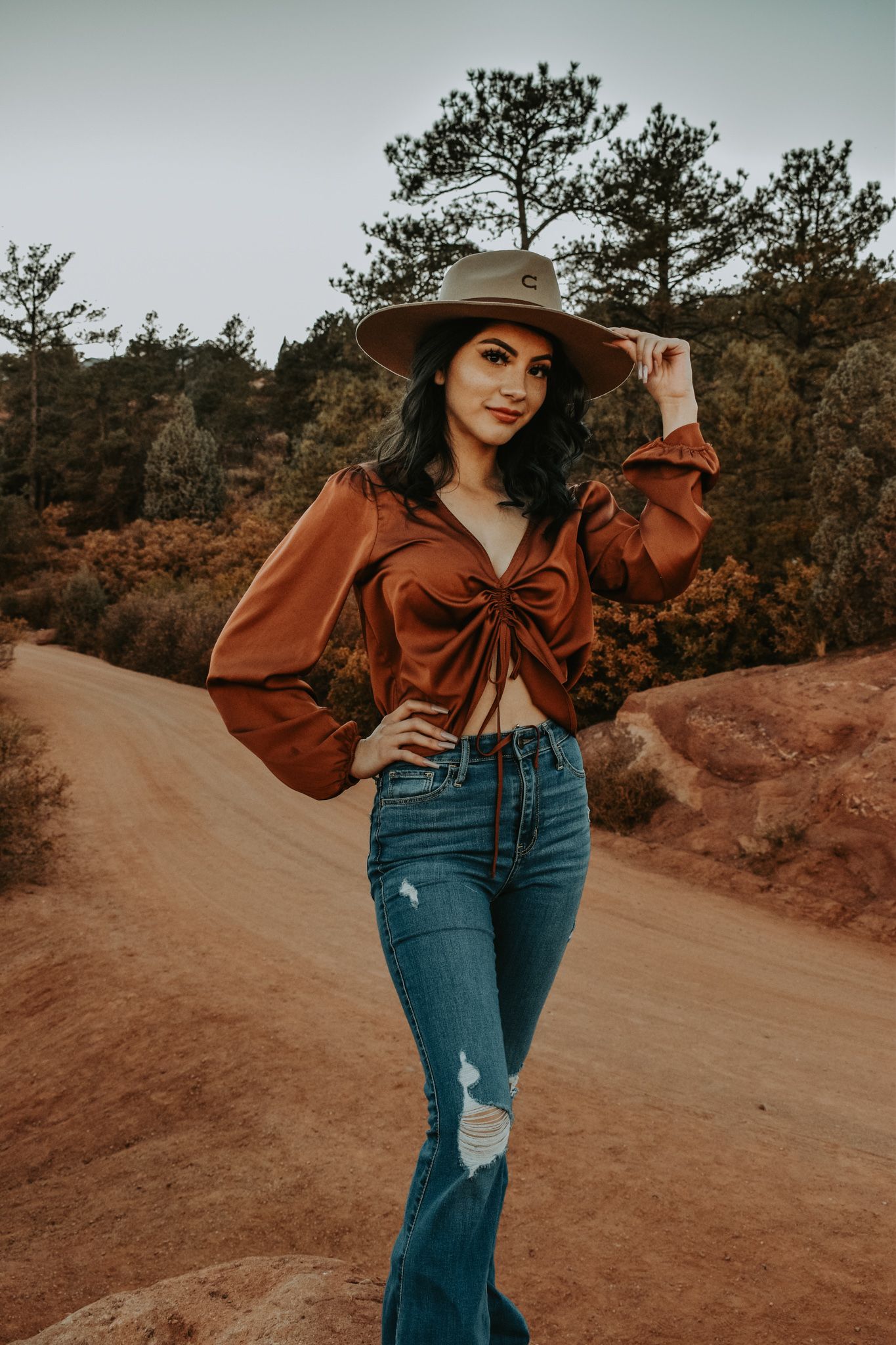 Stylish woman in rust crop top, wide-brim hat, and flare jeans on red dirt road.