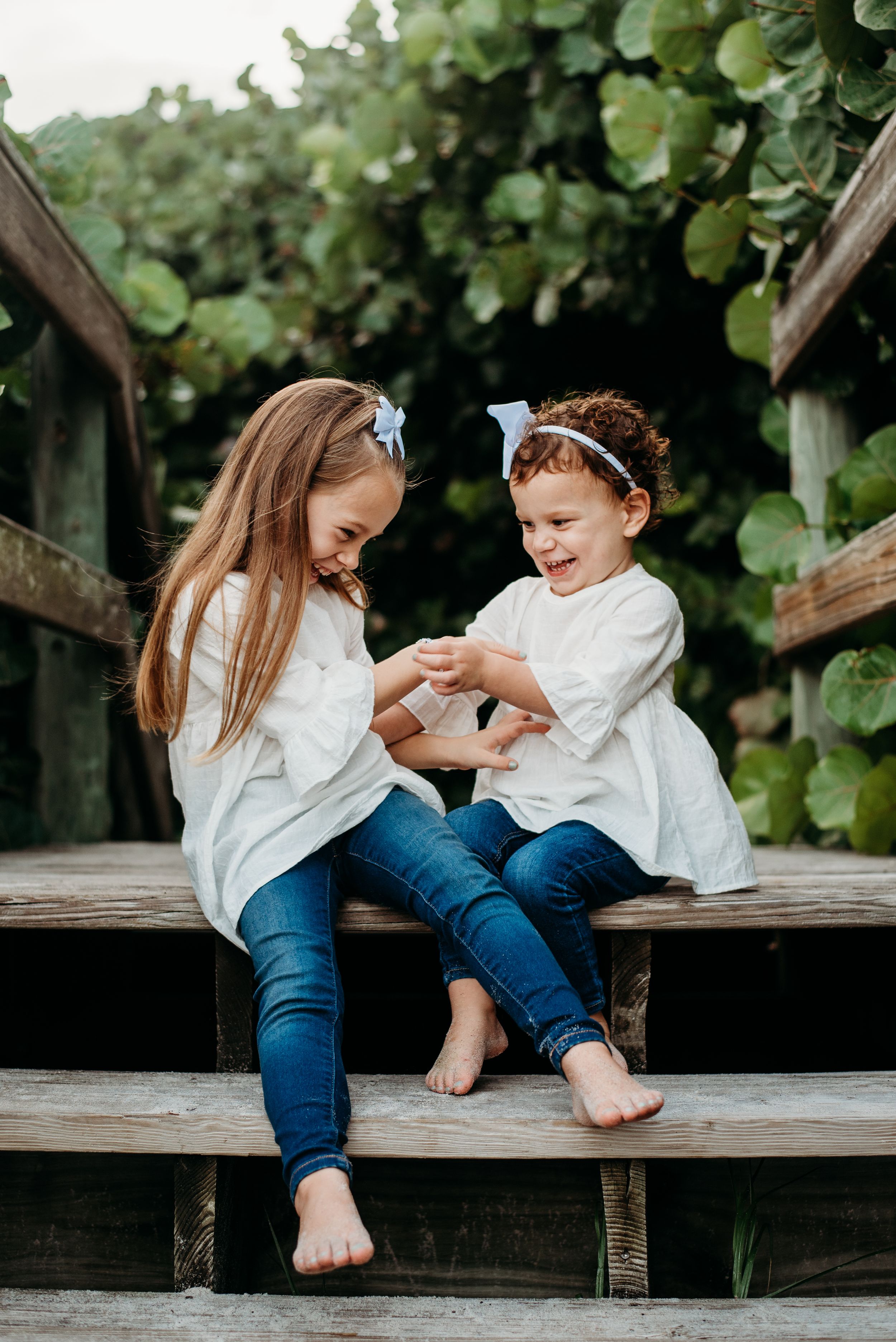 Two children in white shirts sitting together on wooden steps surrounded by greenery.