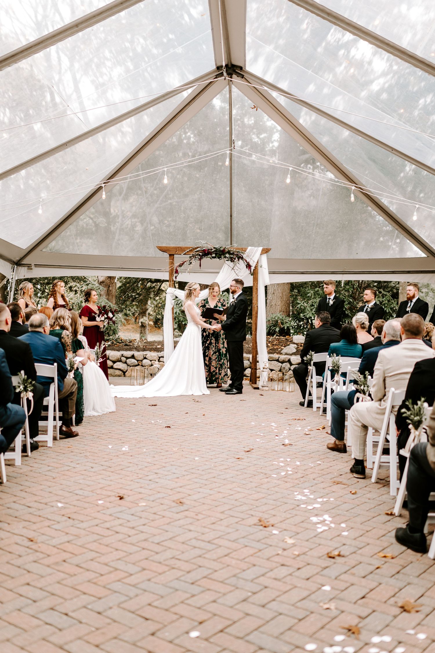 Wide view of wedding ceremony setup under clear tent with brick flooring and white chairs.