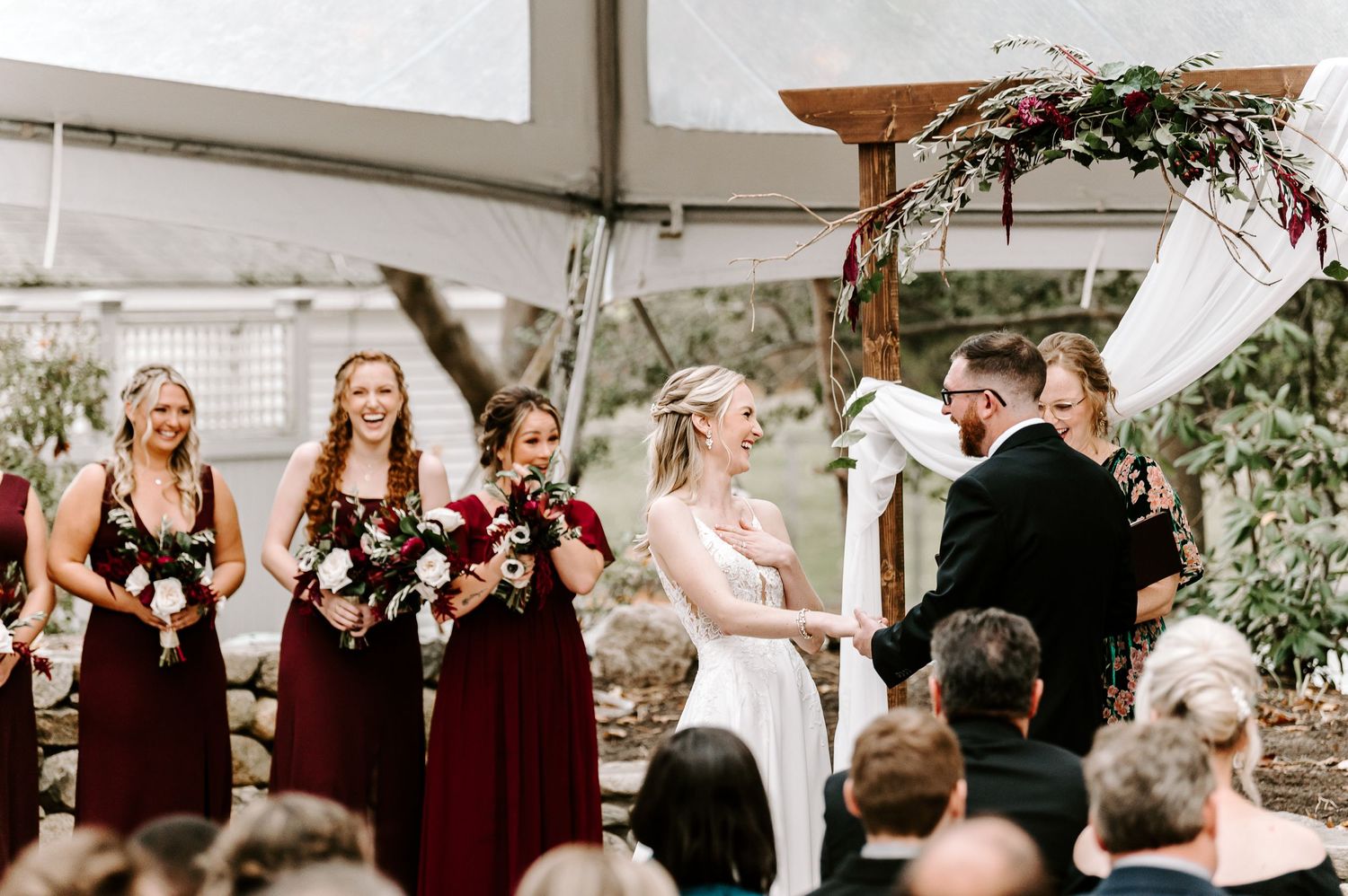 Wedding party celebrating during outdoor ceremony with burgundy and black attire under white tent.
