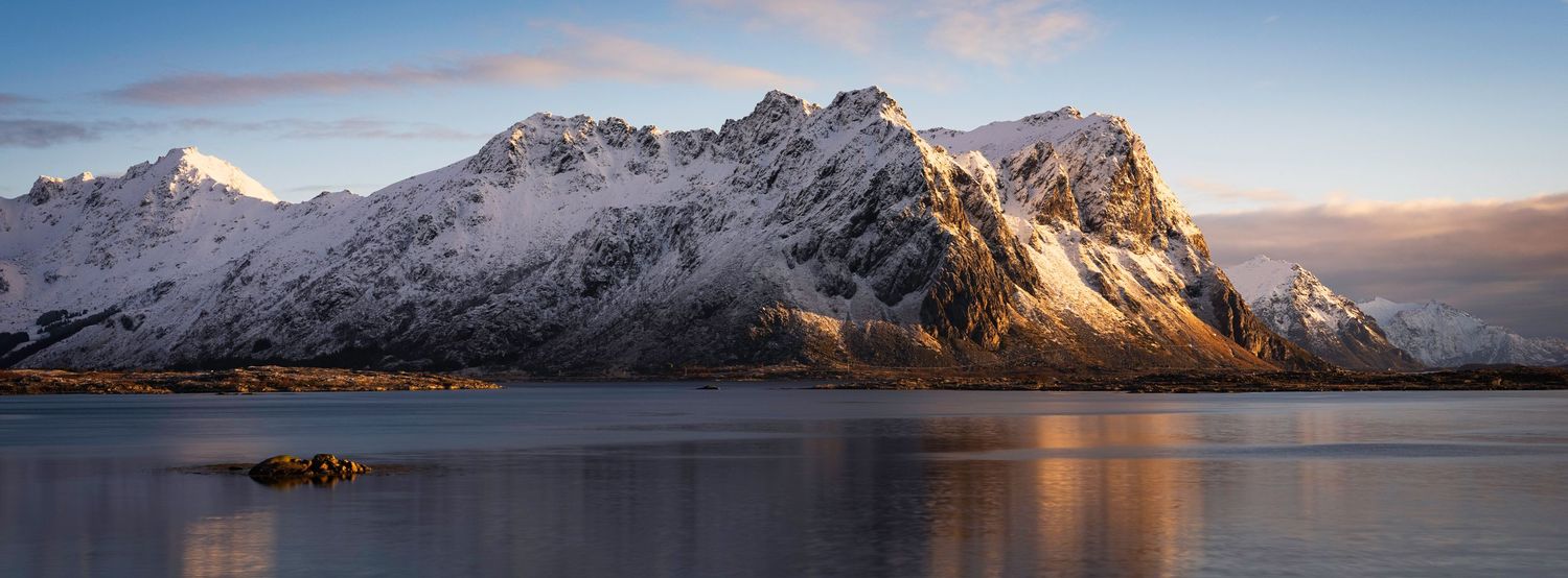 Panoramic view of snow-covered mountain peaks reflecting in calm waters at sunset.