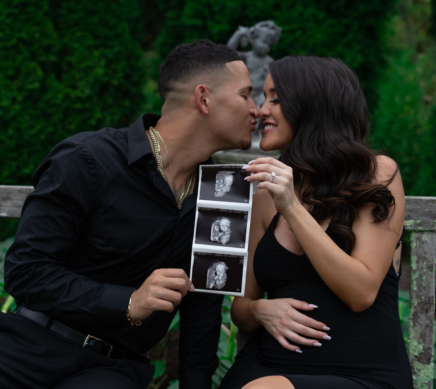 Couple sharing intimate moments while holding ultrasound photos in a garden setting.