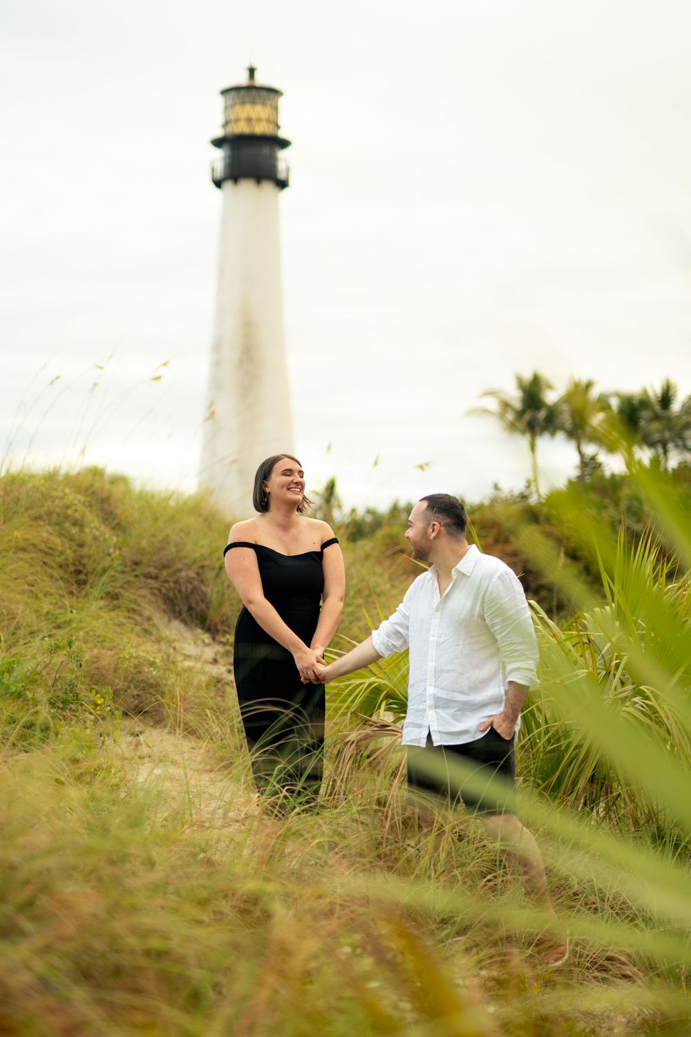 Couple laughs together on grassy hill with lighthouse in background.