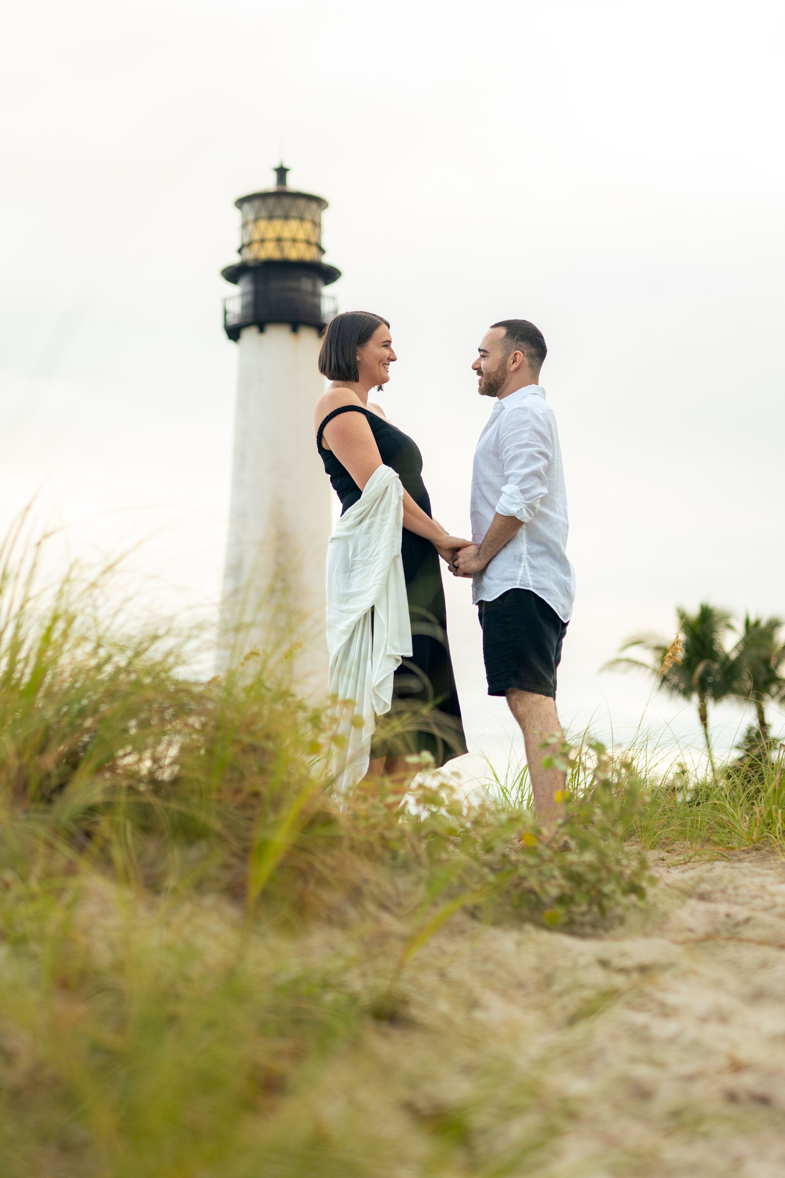 Couple poses near lighthouse with sea grass in foreground.