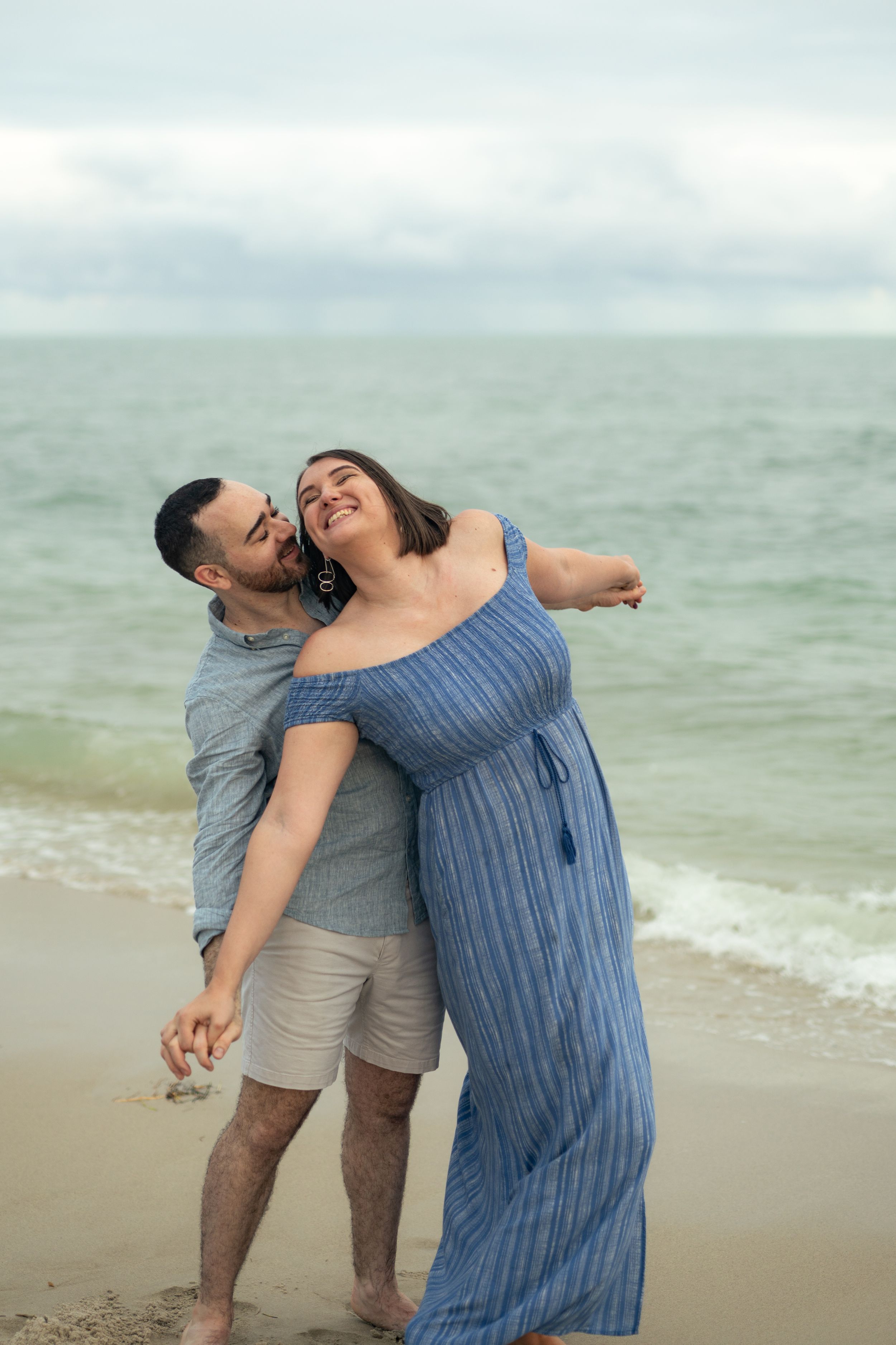 A couple shares a joyful moment on the beach, embracing in casual attire by the ocean.