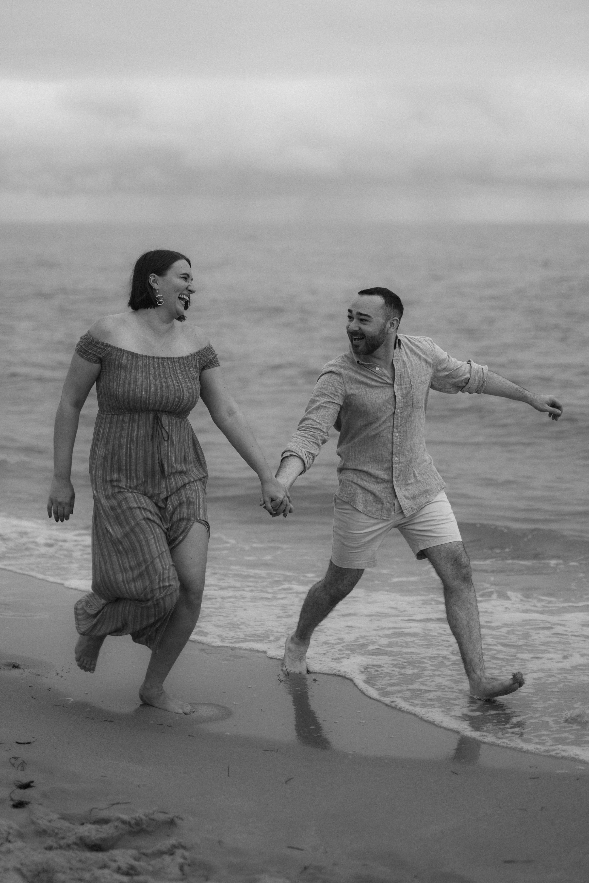 Two people playfully run and dance together along the beach shore in black and white.