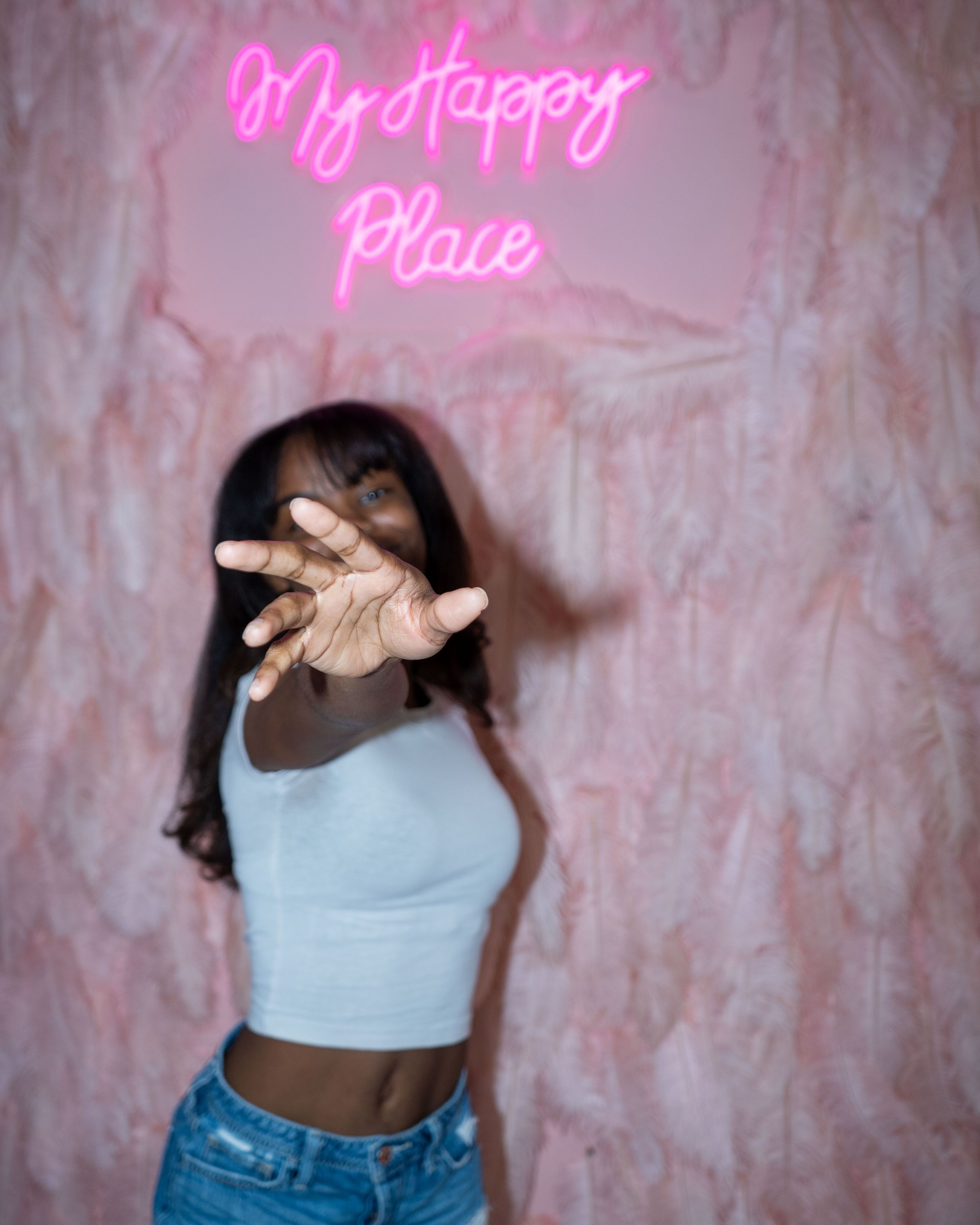 Person posing with hands in front of pink neon sign reading 'My Happy Place'.