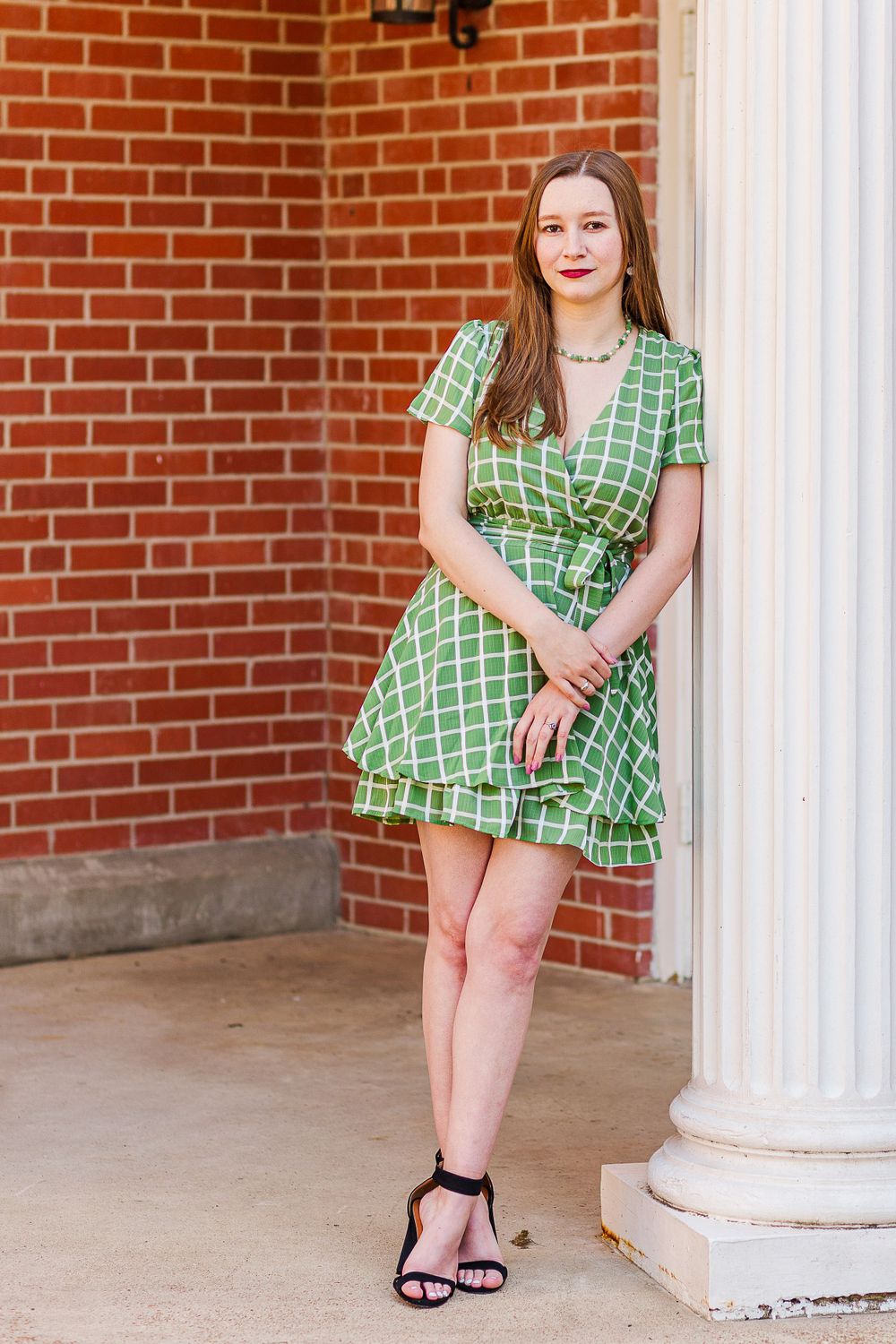 Graduate in green patterned wrap dress standing by white column.