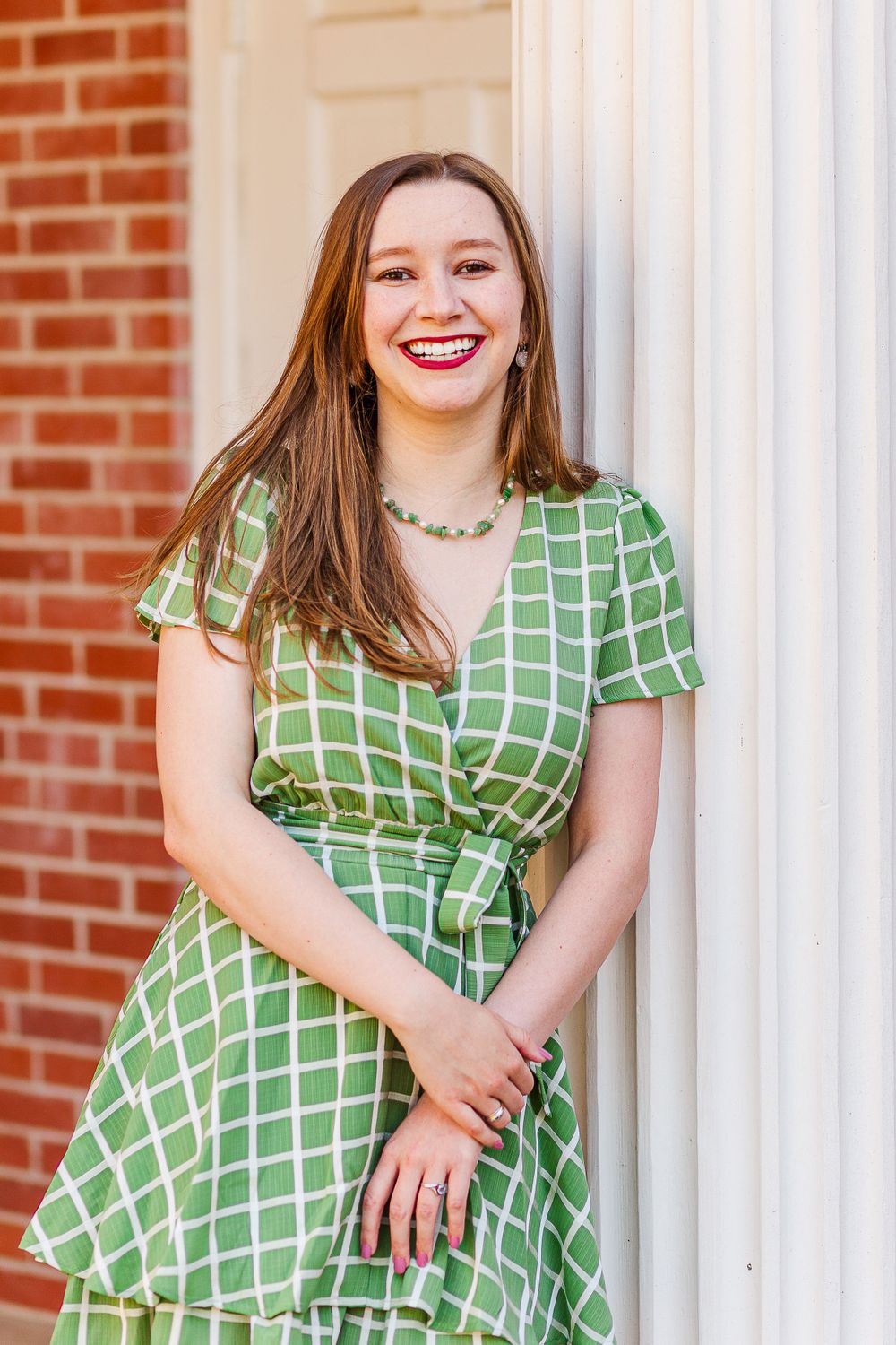 Graduate in green checkered dress smiling by white column.