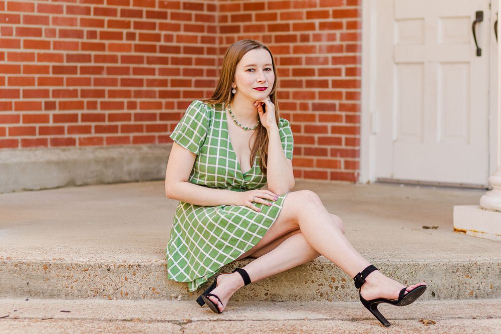 A person in a green checkered dress sits casually on concrete steps with black sandals against a brick wall background.
