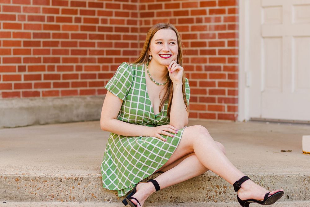 A person in a green plaid dress poses playfully while sitting on steps near a white door and brick wall.