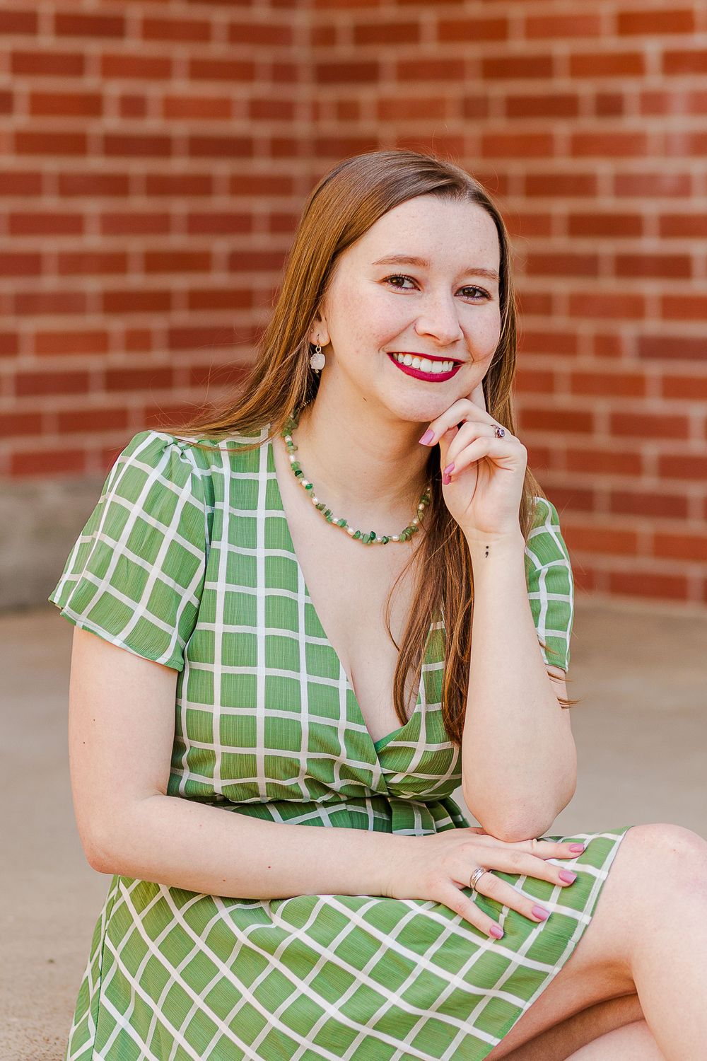 A person wearing a green grid-patterned dress and beaded necklace poses against brick wall background.