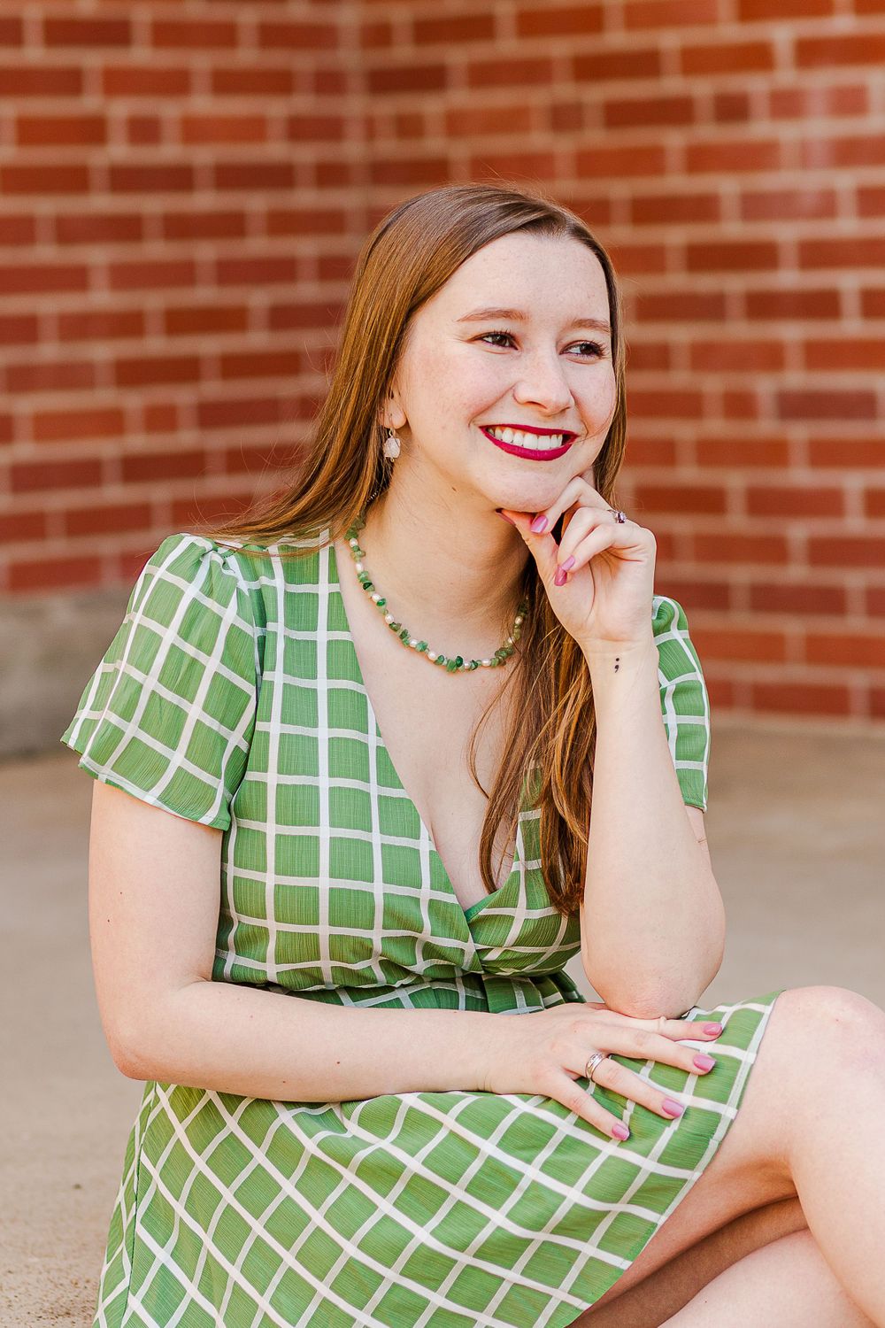 A person in a green checkered wrap dress and necklace smiles while posing against red brick wall.