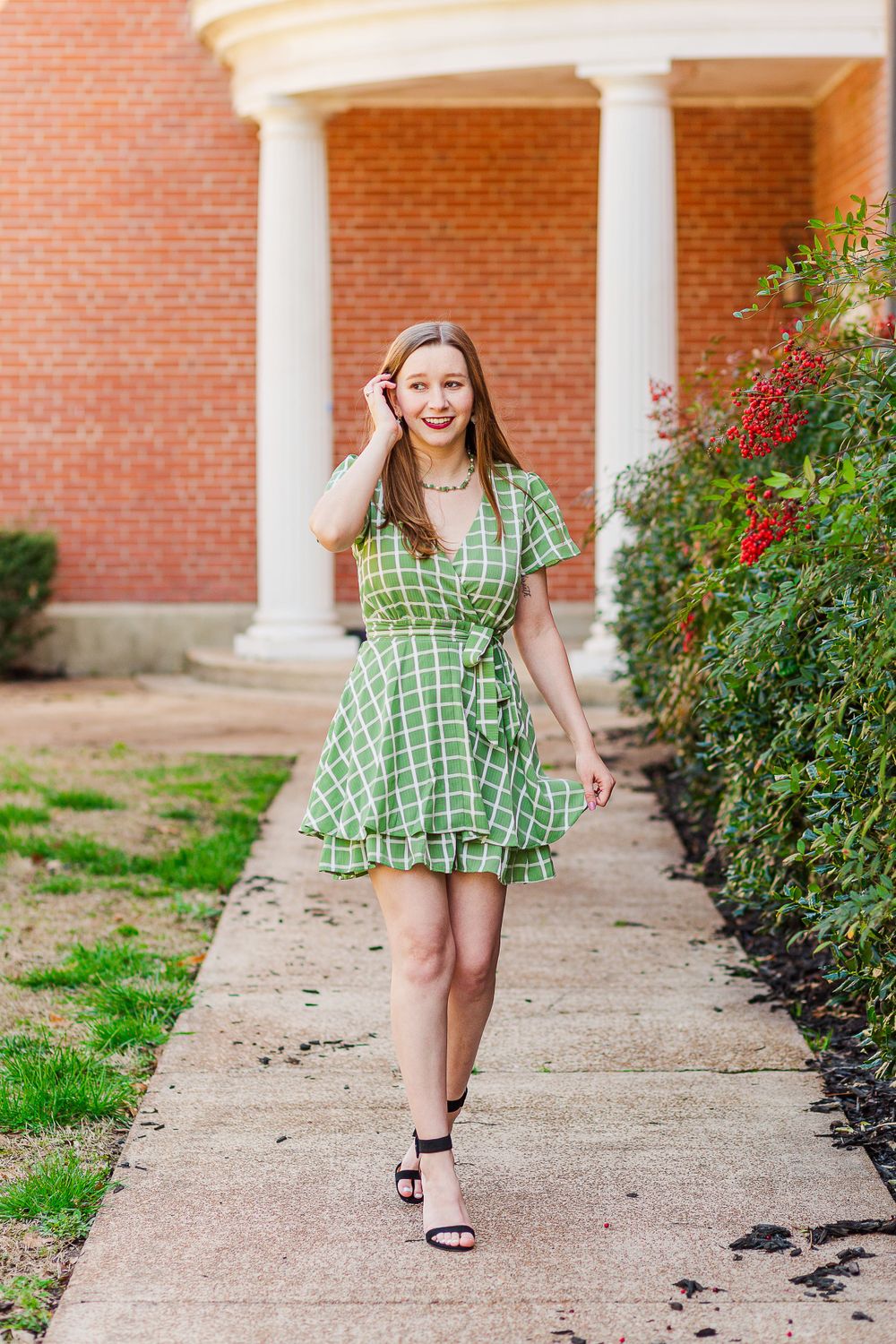 A person in a green plaid dress walks along a pathway with white columns and flowering bushes.
