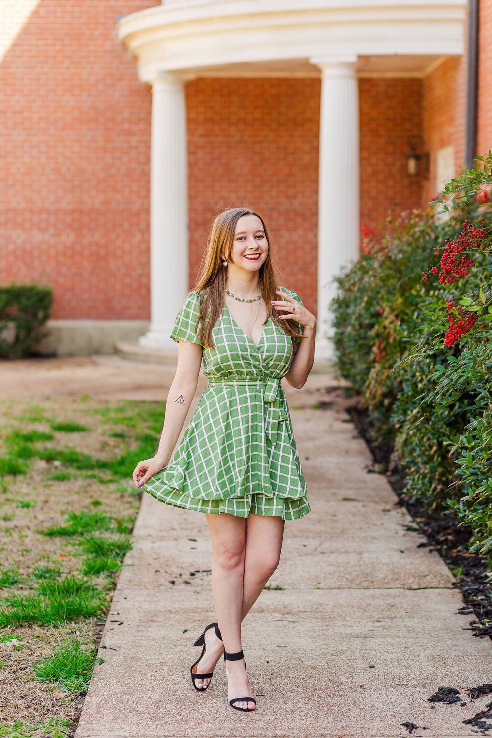A person in a flowy green dress strolls down a walkway with classical columns and red flowers.